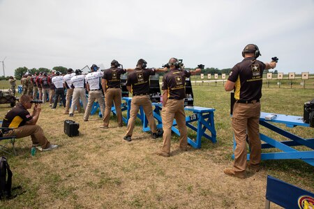 Multiple men in shooting uniforms firing pistol on outdoor pistol range.