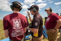 Man in USAMU shooting uniform instructing 2 men on outdoor pistol range.