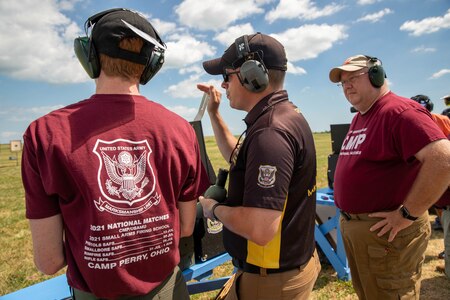 Man in USAMU shooting uniform instructing 2 men on outdoor pistol range.