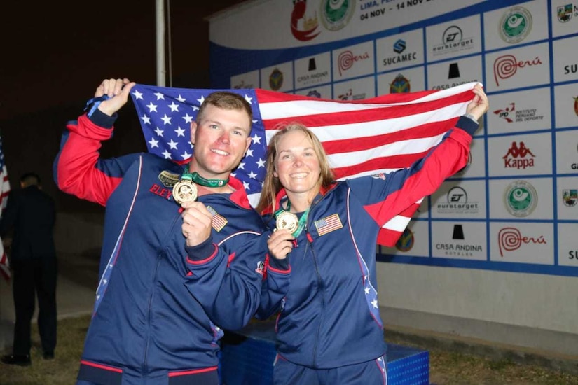 Man and woman in U.S.A. uniform celebrating with medals in front of American flag.