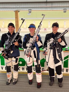 Women in shooting uniforms posing with their air rifles inside at event.