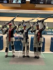 Women in shooting uniforms posing in celebration with their air rifles inside at event.