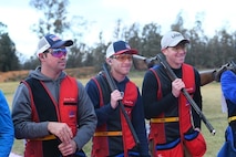 Men in shooting uniforms posing with their shotguns outside at event.