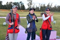 3 women in shooting uniforms posing in celebration with their shotguns outside at event.
