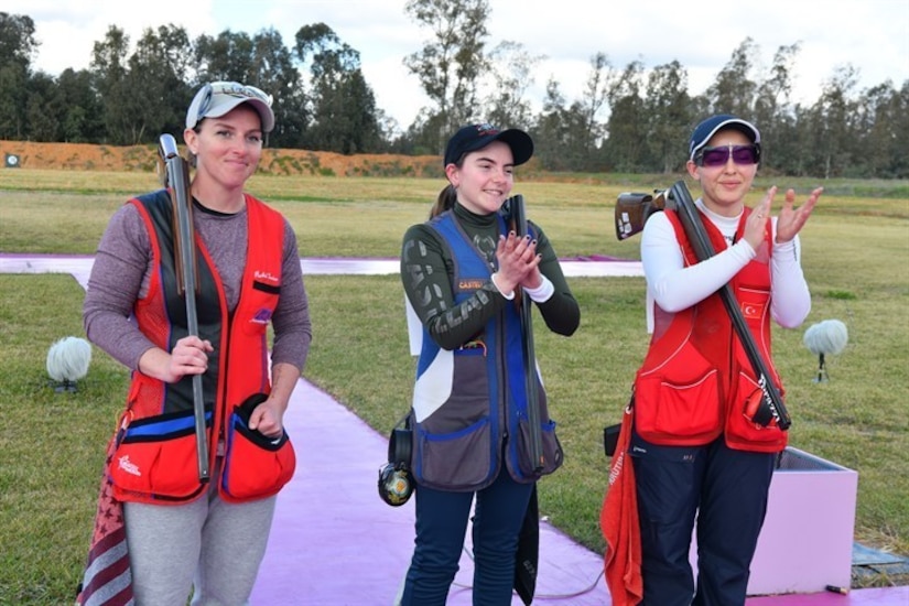 3 women in shooting uniforms posing in celebration with their shotguns outside at event.