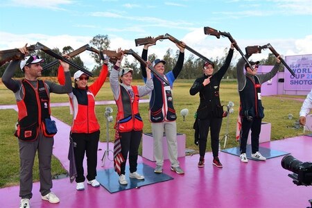 Men and women in shooting uniforms posing in celebration with their shotguns outside at event.