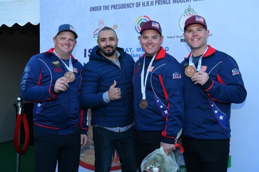 4 men in U.S.A. uniform celebrating with medals in front of event banner.