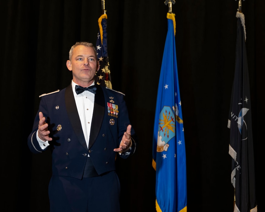 Man in dressed uniform speaks while making hand gestures. Behind him are the United States, Air Force and Space Force flags.