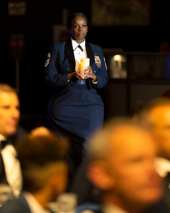 An African-American woman, in mess-dress uniform, carries a candle. In the foreground are out-of-focus diners in yellow light.