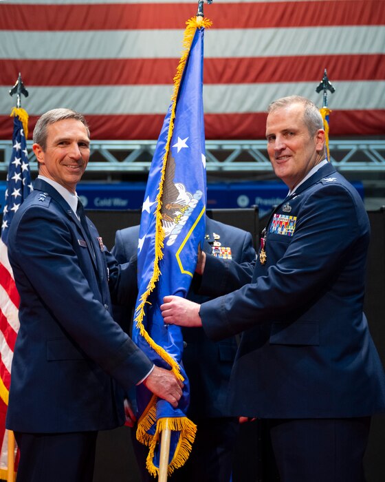 Two men in uniform stand holding a flag between them. Both of them have their heads turned looking at the camera.