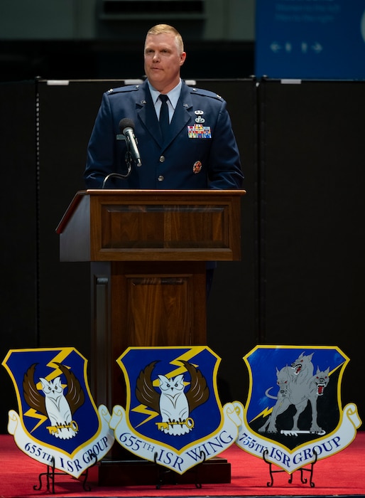 A man in uniform speaks at a podium. In front of the podium are the shields/logos of the 655th ISR Wing, the 655th ISR Group and the 755th ISR Group.