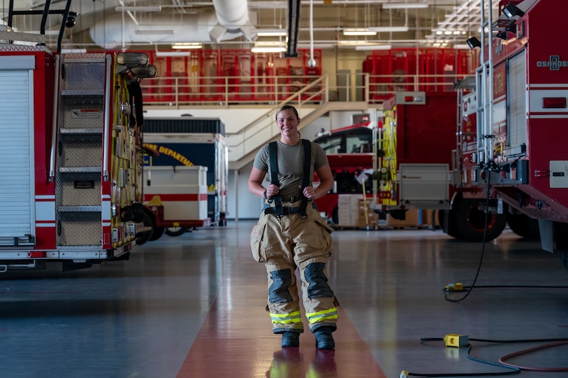 A woman wearing firefighter gear stands in front of fire vehicles.