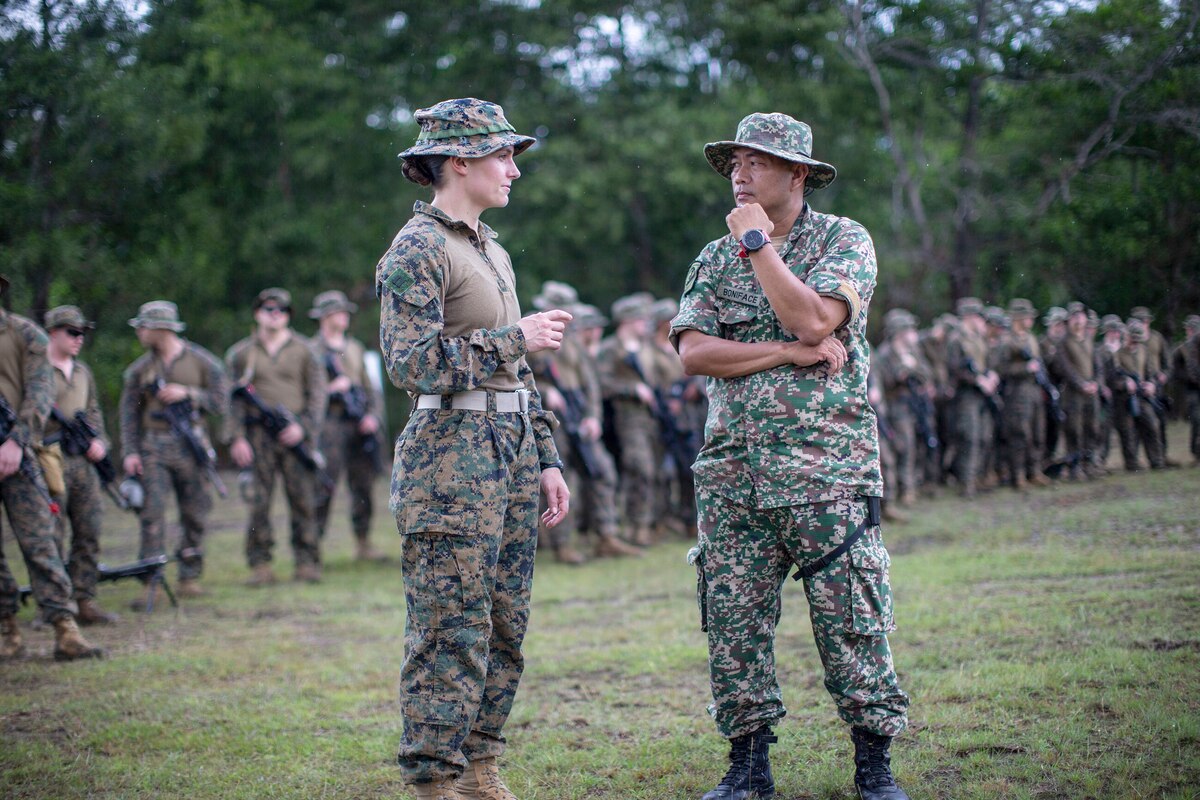 A U.S. Marine talks to a foreign service member on a field as other troops stand by.