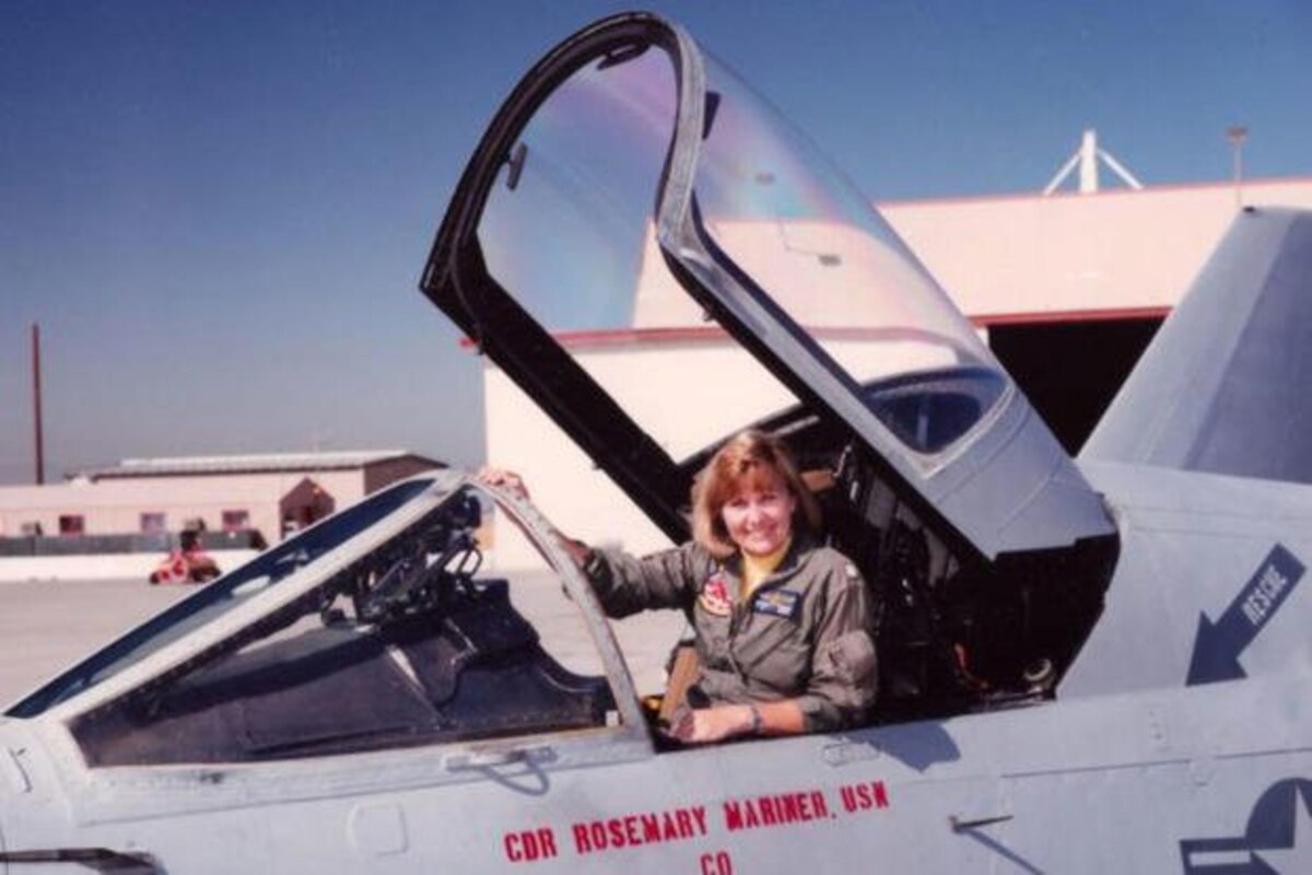 A pilot sits and smiles for a photo in the open cockpit of a parked aircraft.