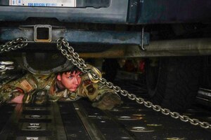 A service member crawls under a truck to ensure security.