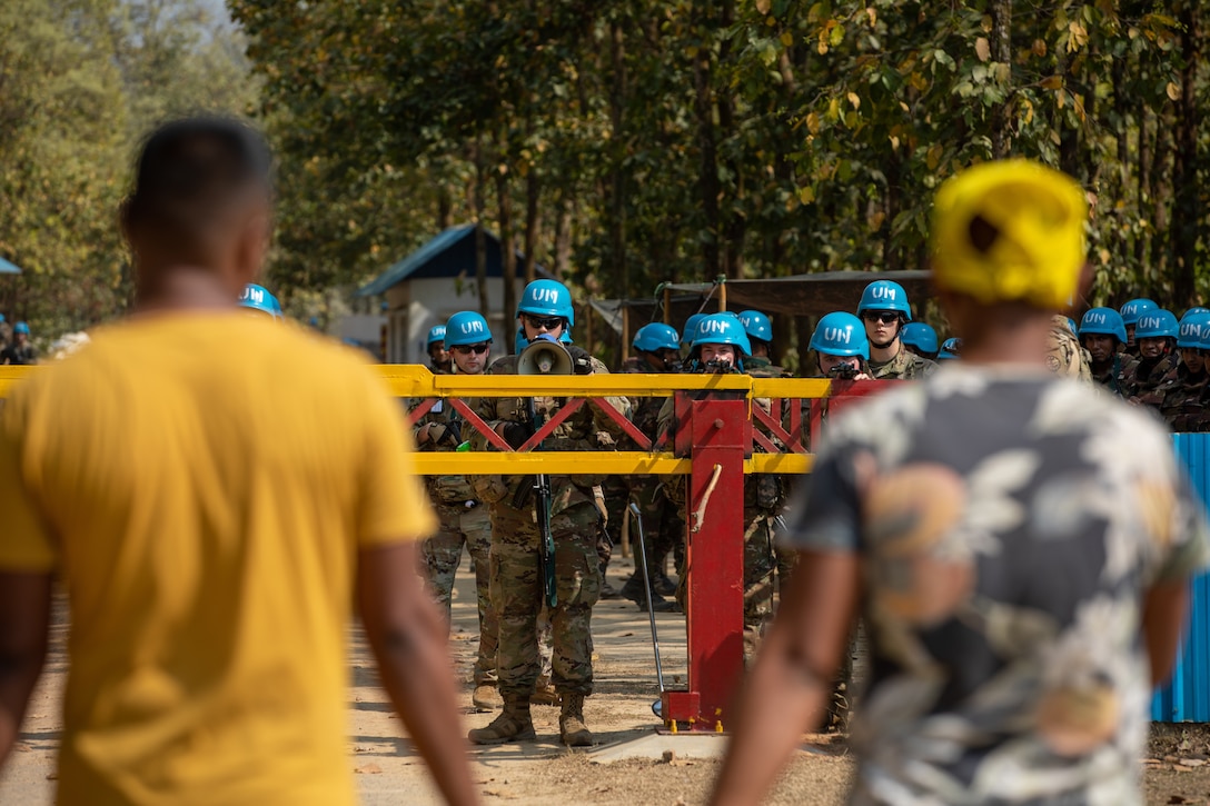 Soldiers with blue helmets stand in a line with weapons as two men walk toward them.