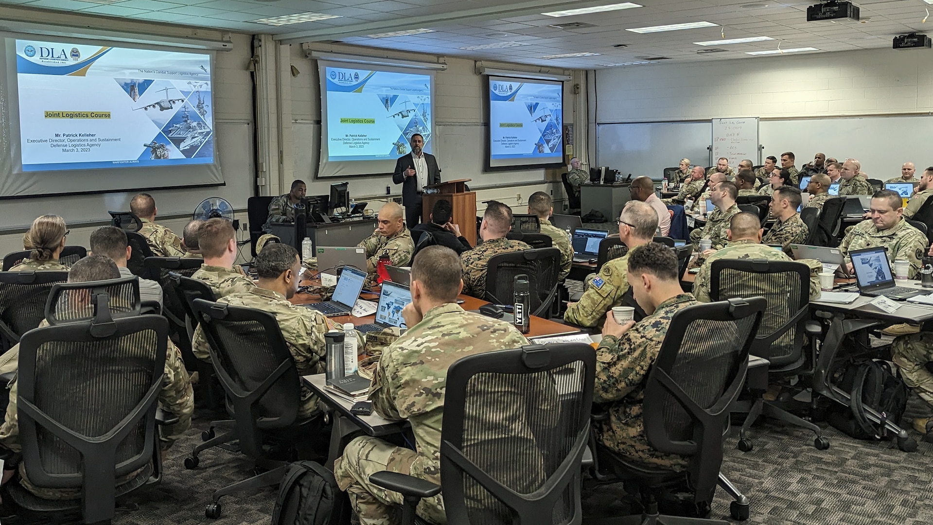 A man with three screens behind him addresses a room full of adult students.