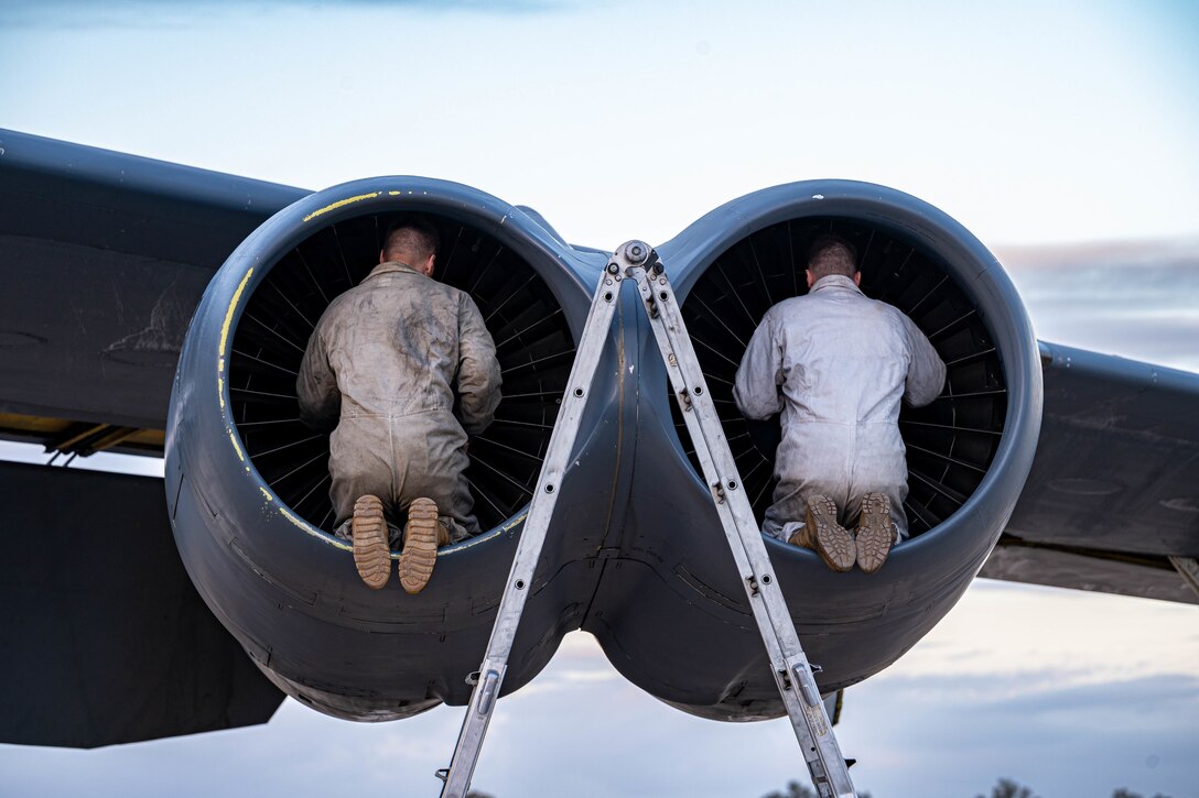 Two airmen kneel inside two large aircraft engines with their backs toward the camera.