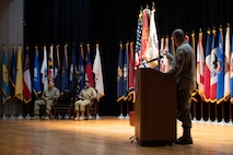 man wearing u.s. army uniform stands behind a podium and points at two people sitting in chairs.