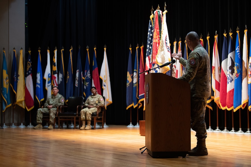 man wearing u.s. army uniform stands behind a podium and points at two people sitting in chairs.