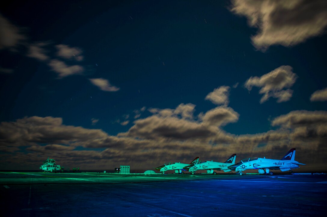 Aircraft illuminated by blue light sit parked on the deck of a ship at night.