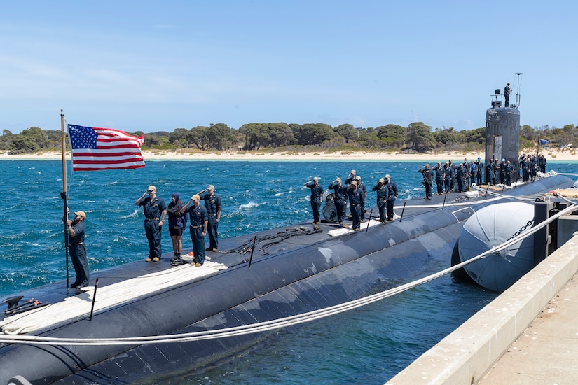 Sailors salute a U.S. flag atop a submarine moored at a pier.