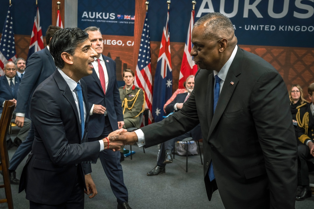 Secretary of Defense Lloyd J. Austin III shakes hands with the British prime minister in a room with other officials.