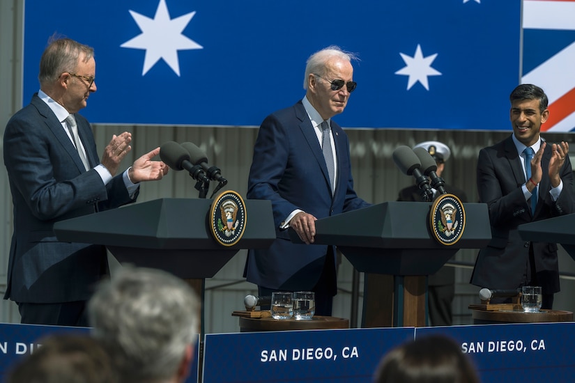 The British and Australian prime ministers applaud for President Joe Biden as they all stand at lecterns.