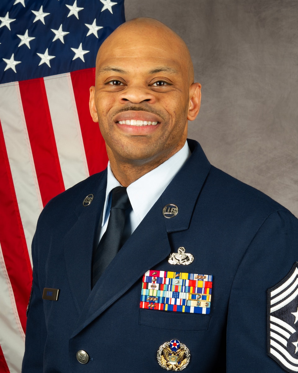 African American man in blue uniform with American flag behind his right shoulder.