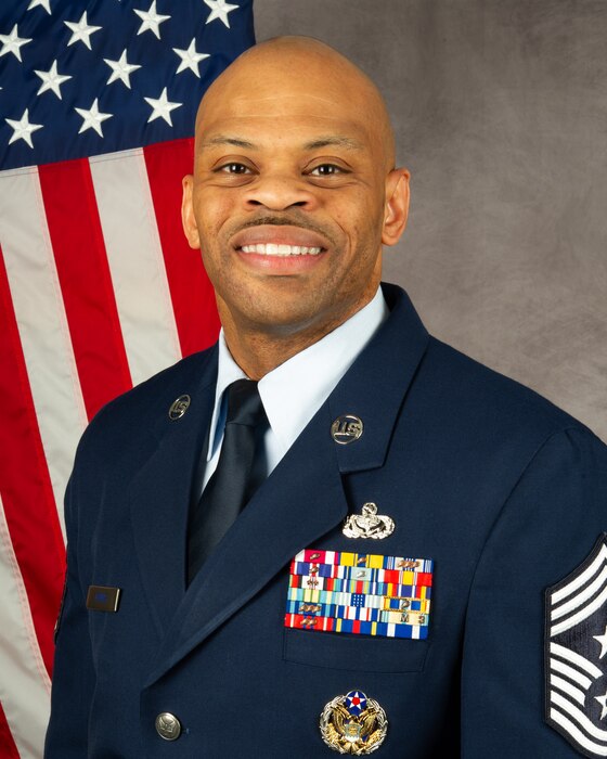 African American man in blue uniform with American flag behind his right shoulder.