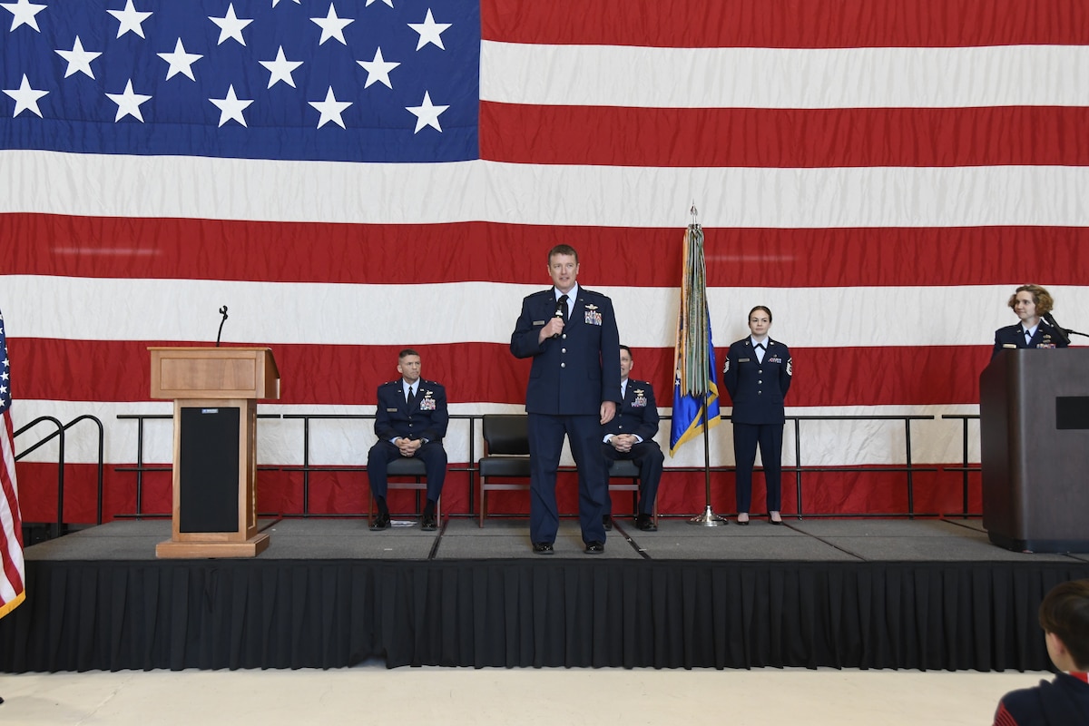 Service man speaks on stage in front of large American flag. Four other service members are on stage behind man speaking, two are sitting and two are standing.
