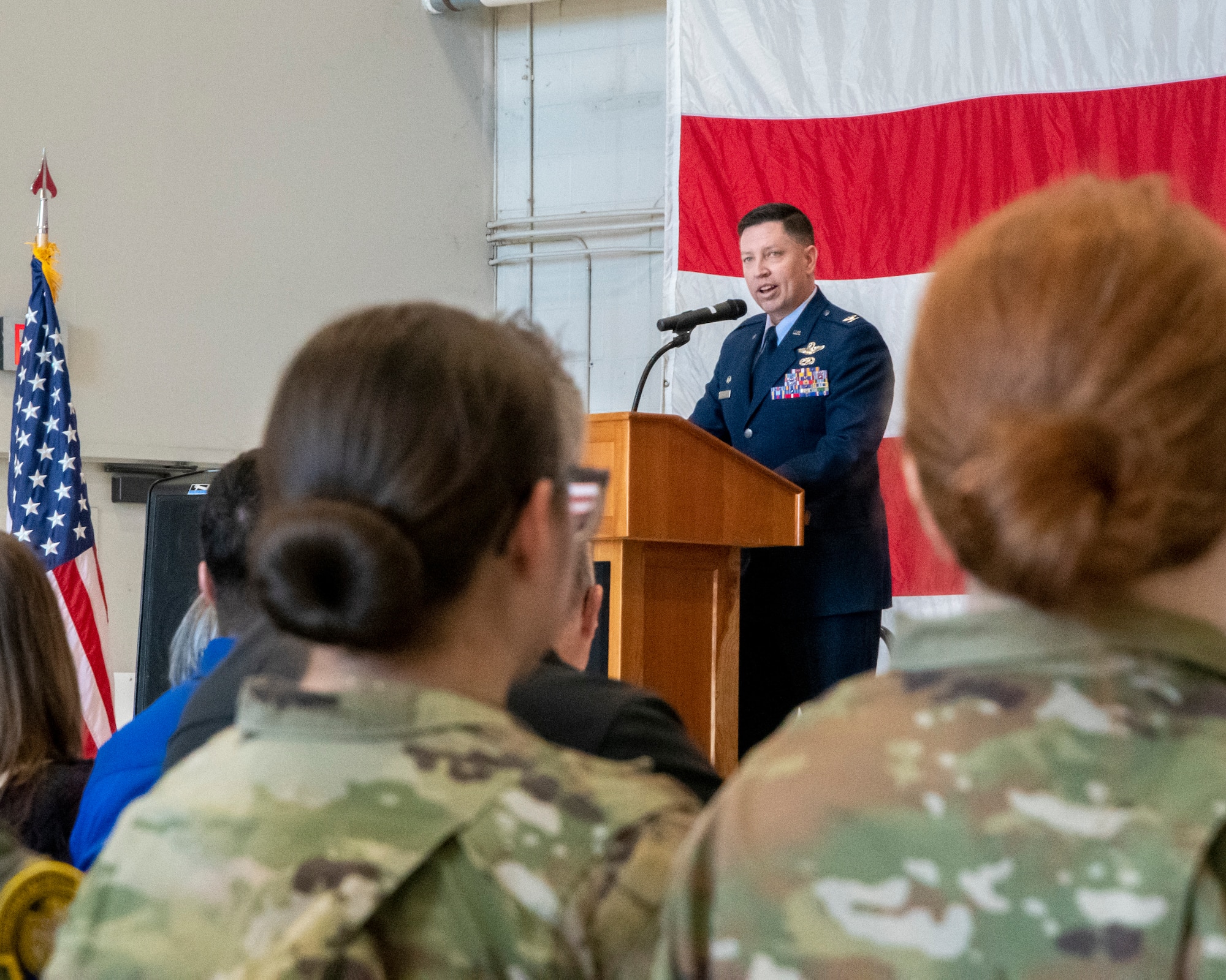 Military member speaks on stage in front of a podium. Picture is taken from the crowd behind two female military members.