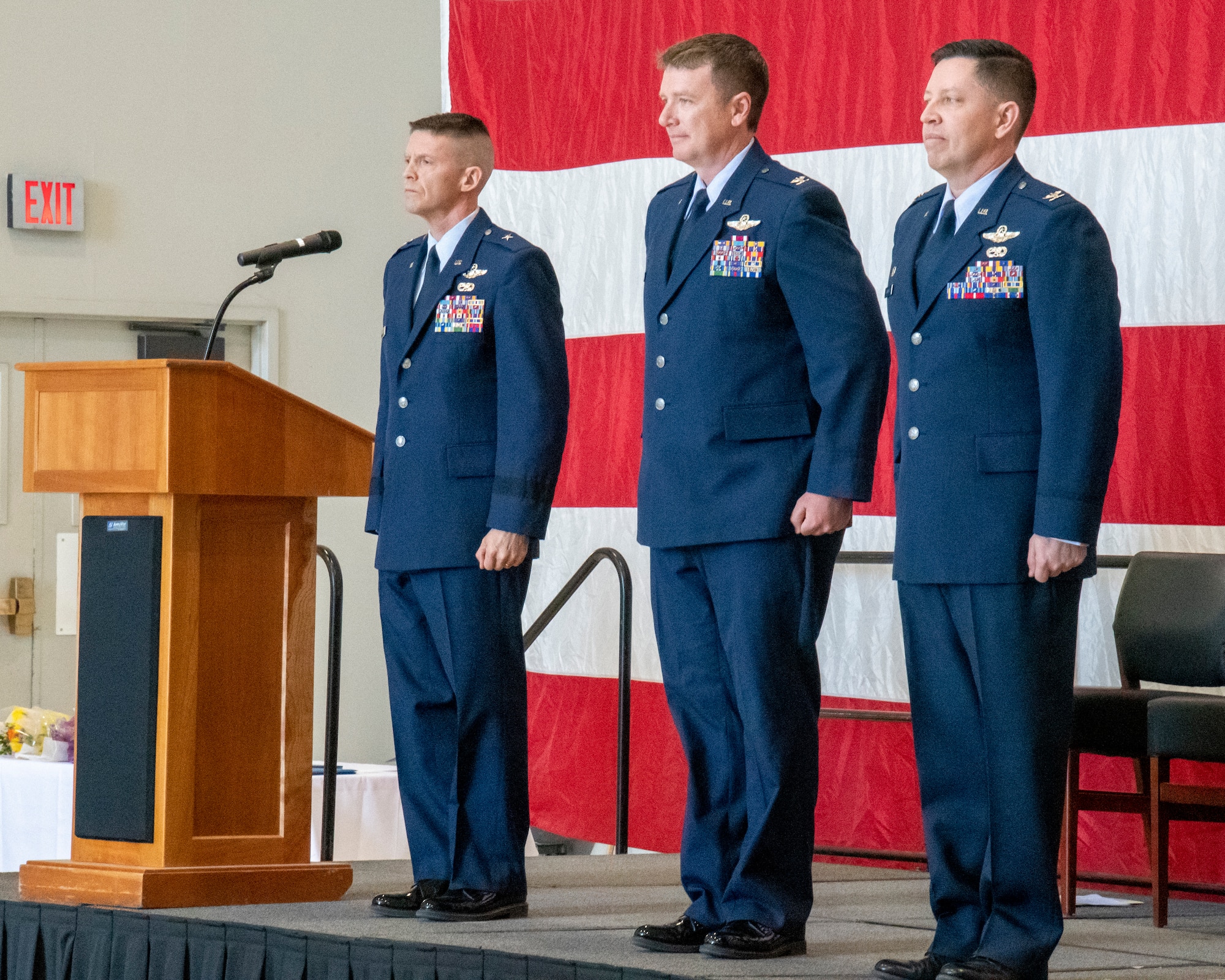 Three men stand side by side on stage at attention in front of large American flag.