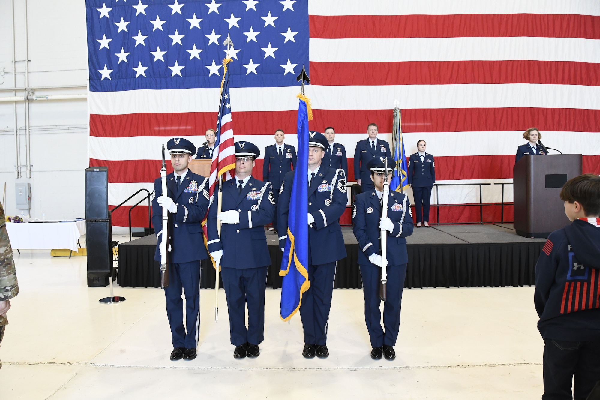 Four honor guard members stand at attention on the ground holding flags and riffles. Behind them is five service members on a stage in front of a large American flag.