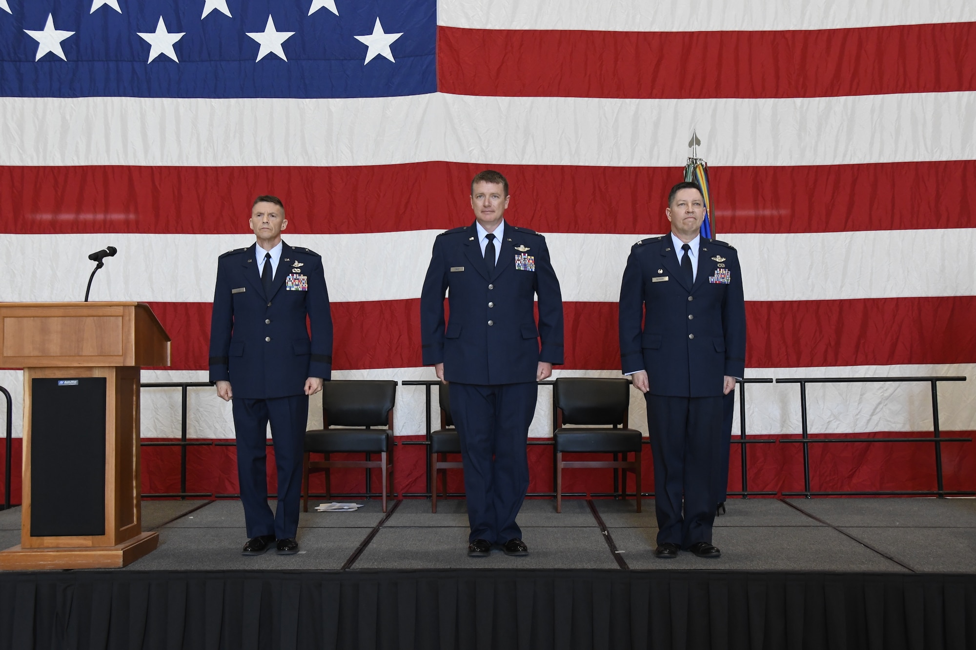 Three service members stand on stage at attention in front of large American flag.