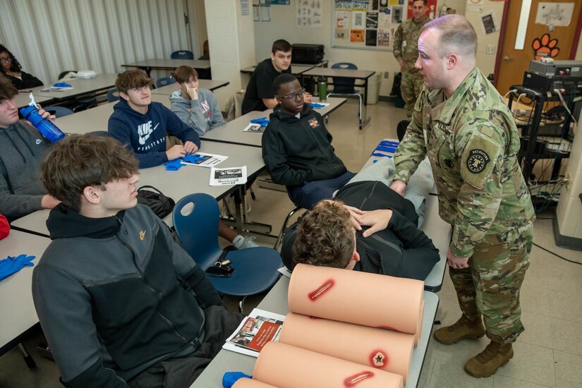 Male Army Soldier demonstrates how to stop bleeding to a school of high school students.
