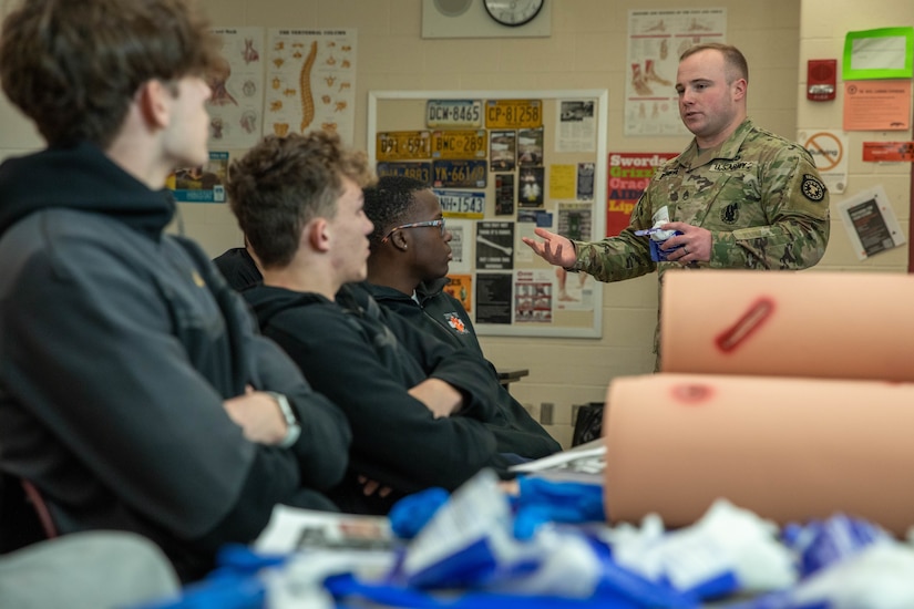Three male high school students listen to male Army Soldier give first aid class.