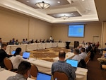 Many individuals seated in a big meeting room in a square configuration looking at a projected slide on a large screen.