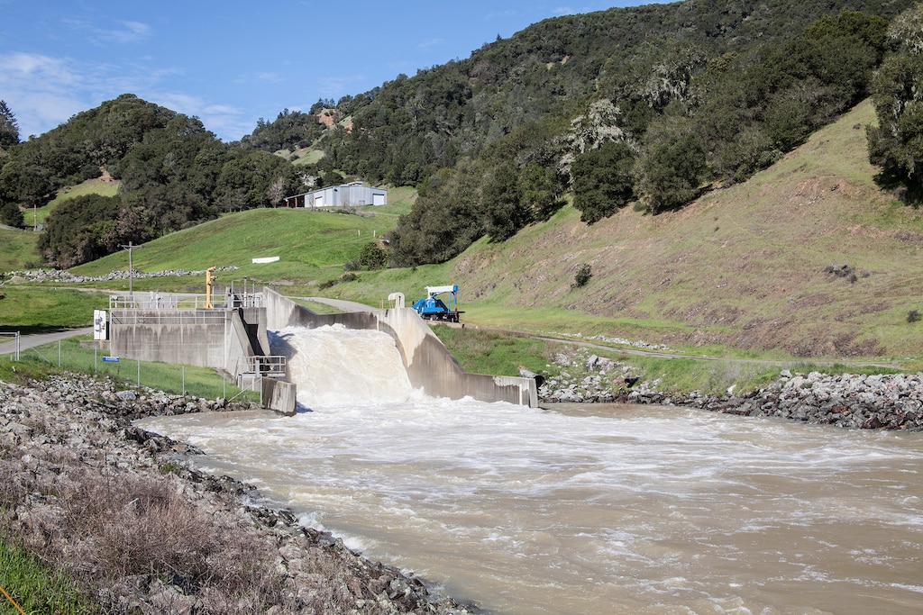 Concrete waterway has lots of water rushing over it with hills and a building in the background.