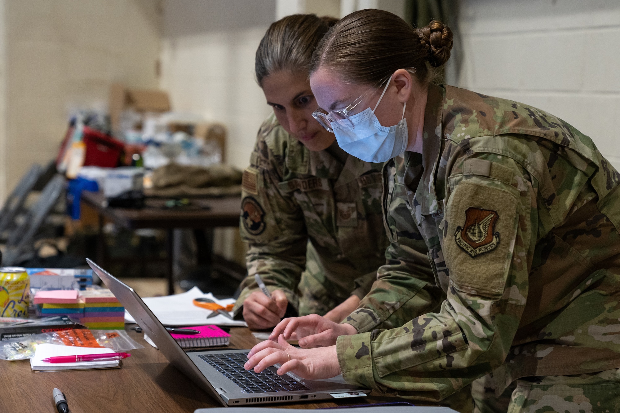 U.S. Air Force Staff Sgt. Zoie King leans over a computer typing while Tech. Sgt. Alexandra Sanders leans over next to her also looking at the computer screen
