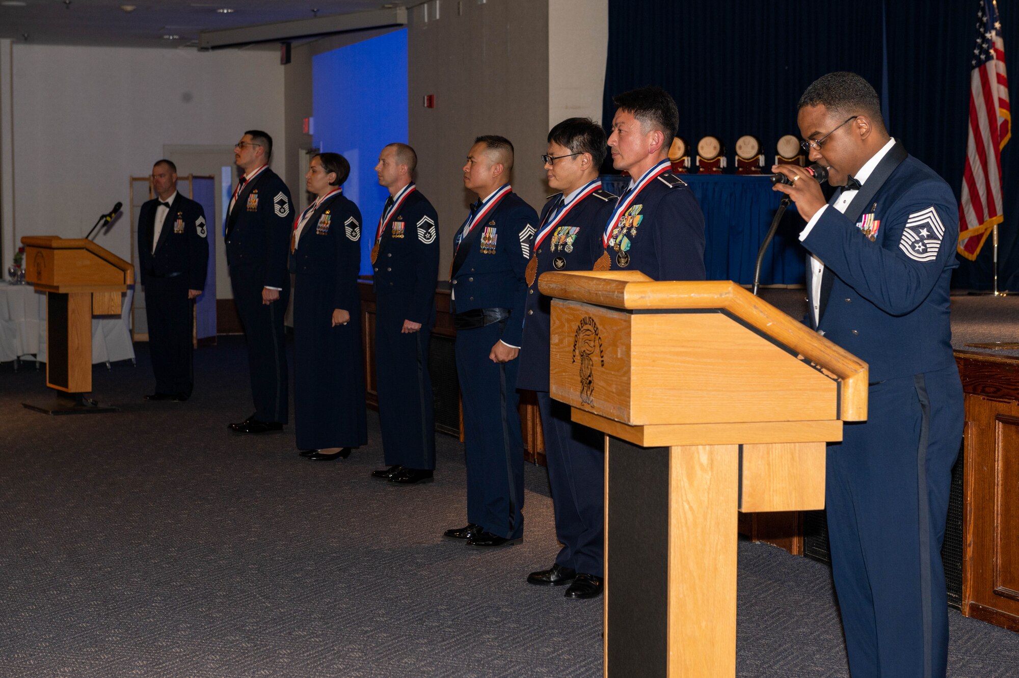 A black man wearing dress blues delivers a speech behind a podium.
