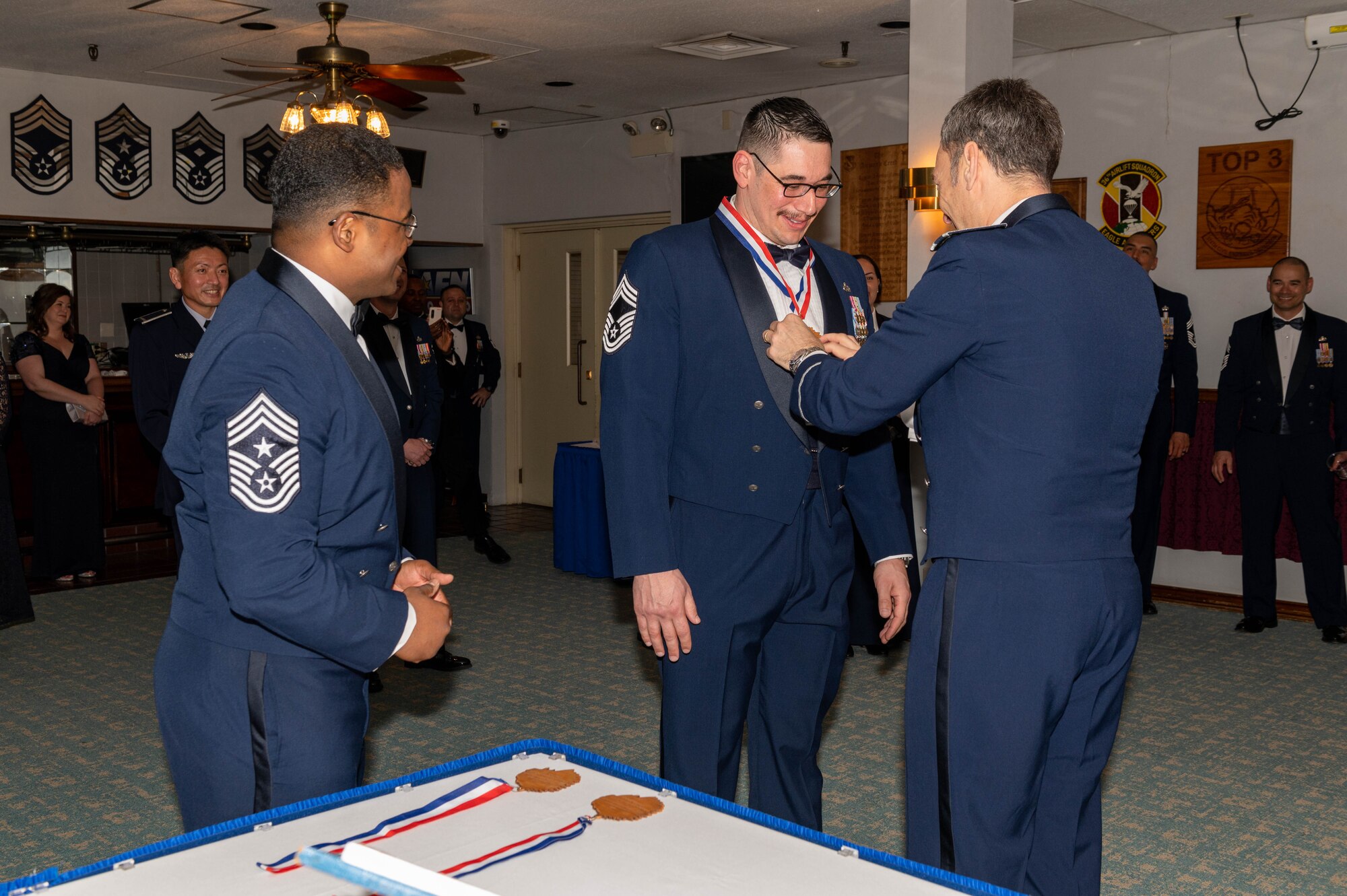 A man wearing dress blues receives a medallion from two other men wearing dress blues.