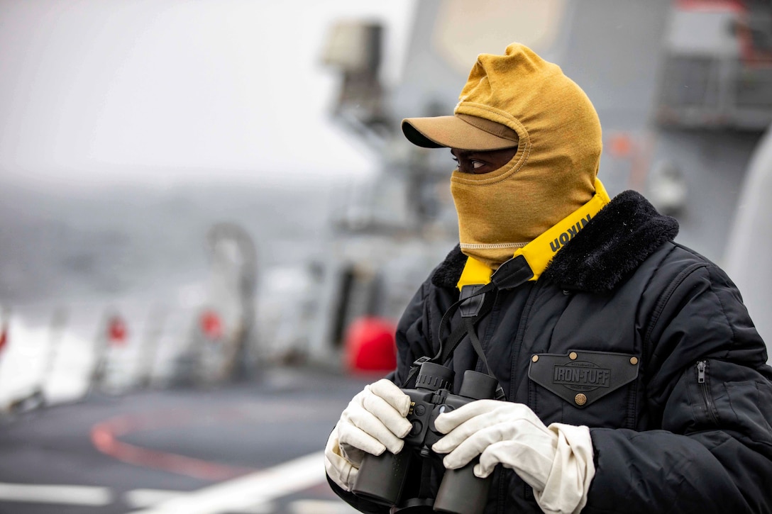 A sailor wearing cold weather gear and gloves holds binoculars on a ship.