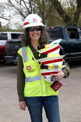 A woman stands in a parking lot in a hardhat and safety vest, and smiles for the camera while holding three hardhats in her left hand.