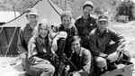 One woman and five men pose for a photo in front of an Army tent.