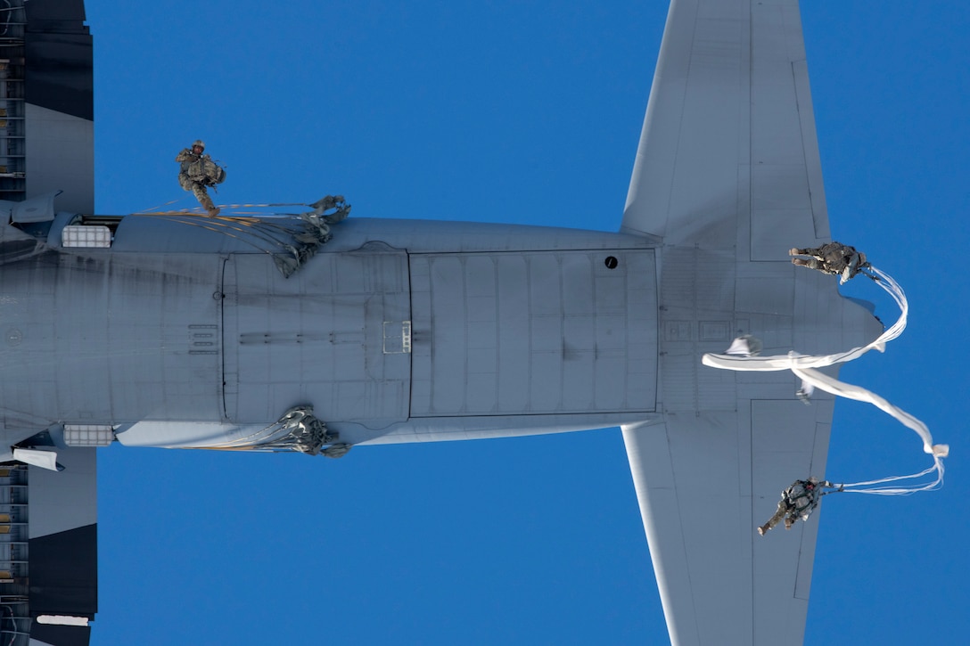 Paratroopers jump out of a large aircraft against a blue sky.