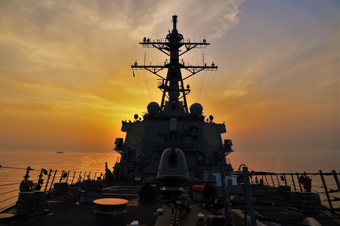 A Navy ship sails toward the camera, illuminated by a low sun in the background.