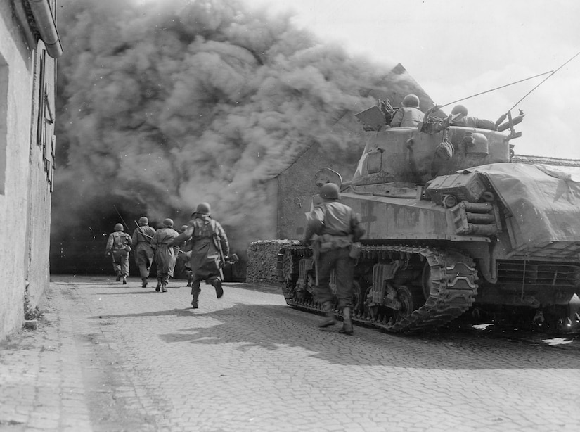 Half a dozen men run toward a smoke plume beside a tank.