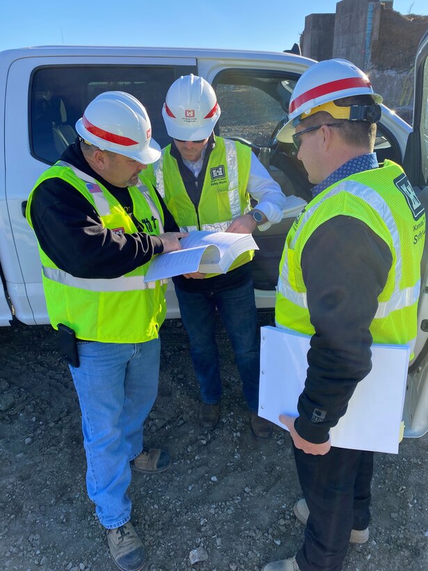Three men in reflective vests and hard hats look at a document in front of a white pick up truck.