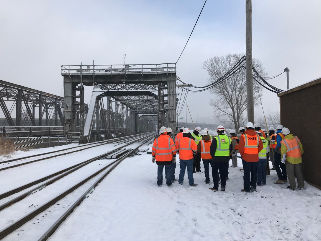 A group of people in reflective vests and hard hats stand in the snow in front of a large grey bridge outside.